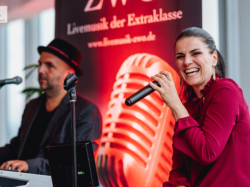 Lachende Sängerin mit Mikrofon, Pianist im Hintergrund, vor Banner "ZWO Livemusik der Extraklasse"