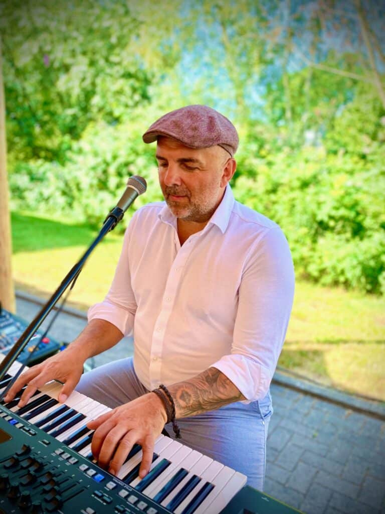 Musiker am Keyboard im Freien, trägt weiße Bluse und Mütze, grüne Natur im Hintergrund.